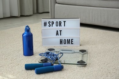 Photo of Fitness equipment and lightbox with hashtag SPORT AT HOME on floor indoors. Message to promote self-isolation during COVID‑19 pandemic