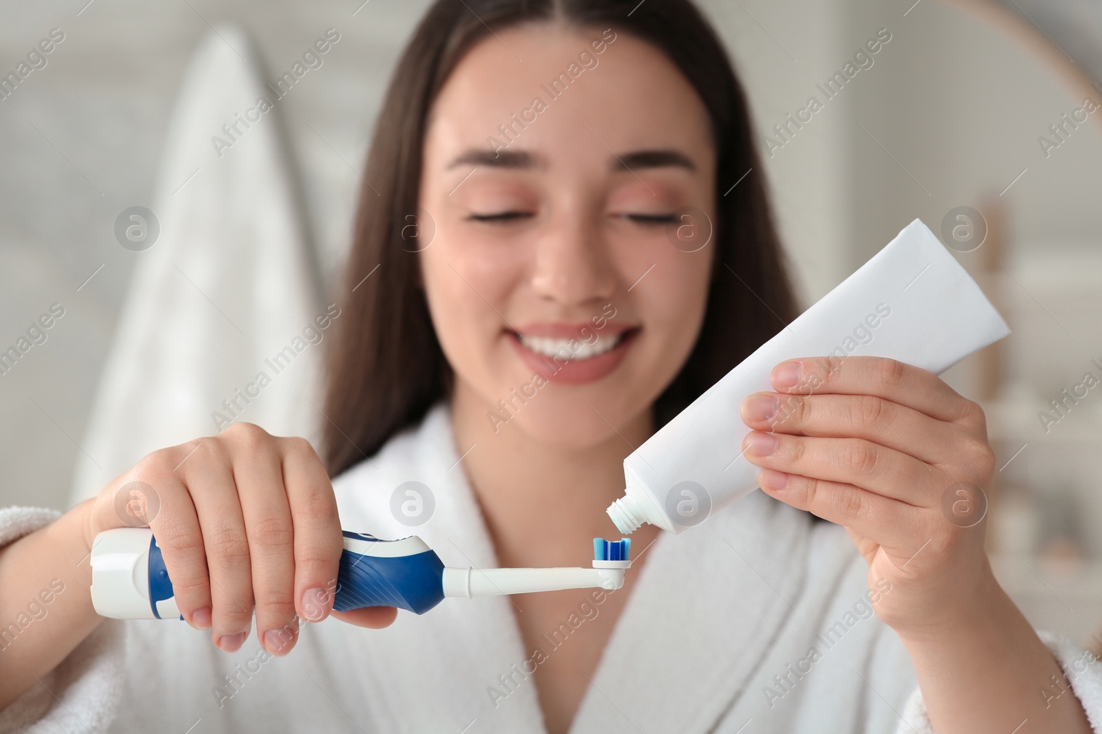 Photo of Young woman squeezing toothpaste from tube onto electric toothbrush in bathroom, selective focus