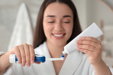 Young woman squeezing toothpaste from tube onto electric toothbrush in bathroom, selective focus