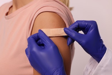 Nurse sticking adhesive bandage on woman's arm after vaccination on light background, closeup