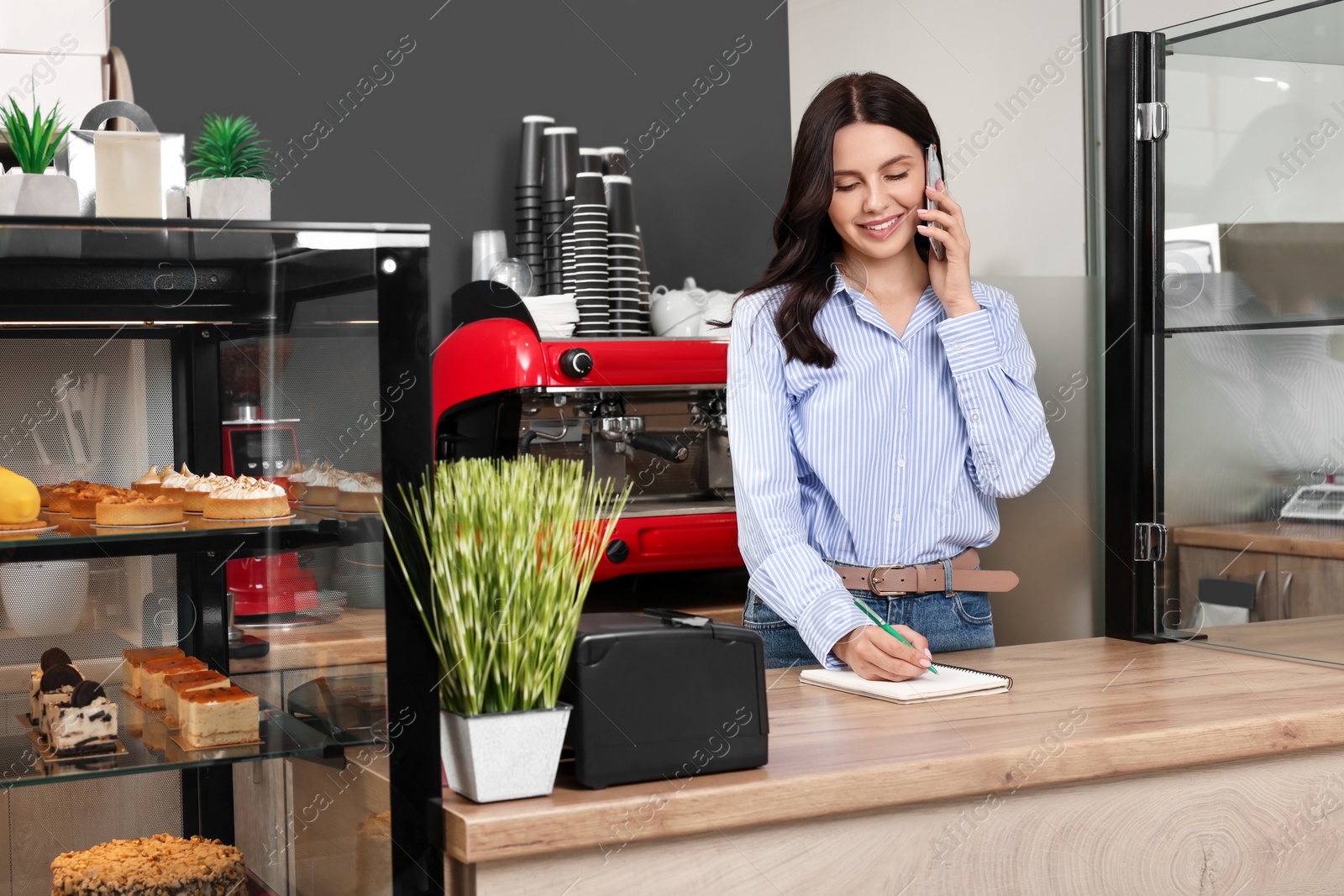 Photo of Business owner talking on phone while working near showcase with desserts in her cafe