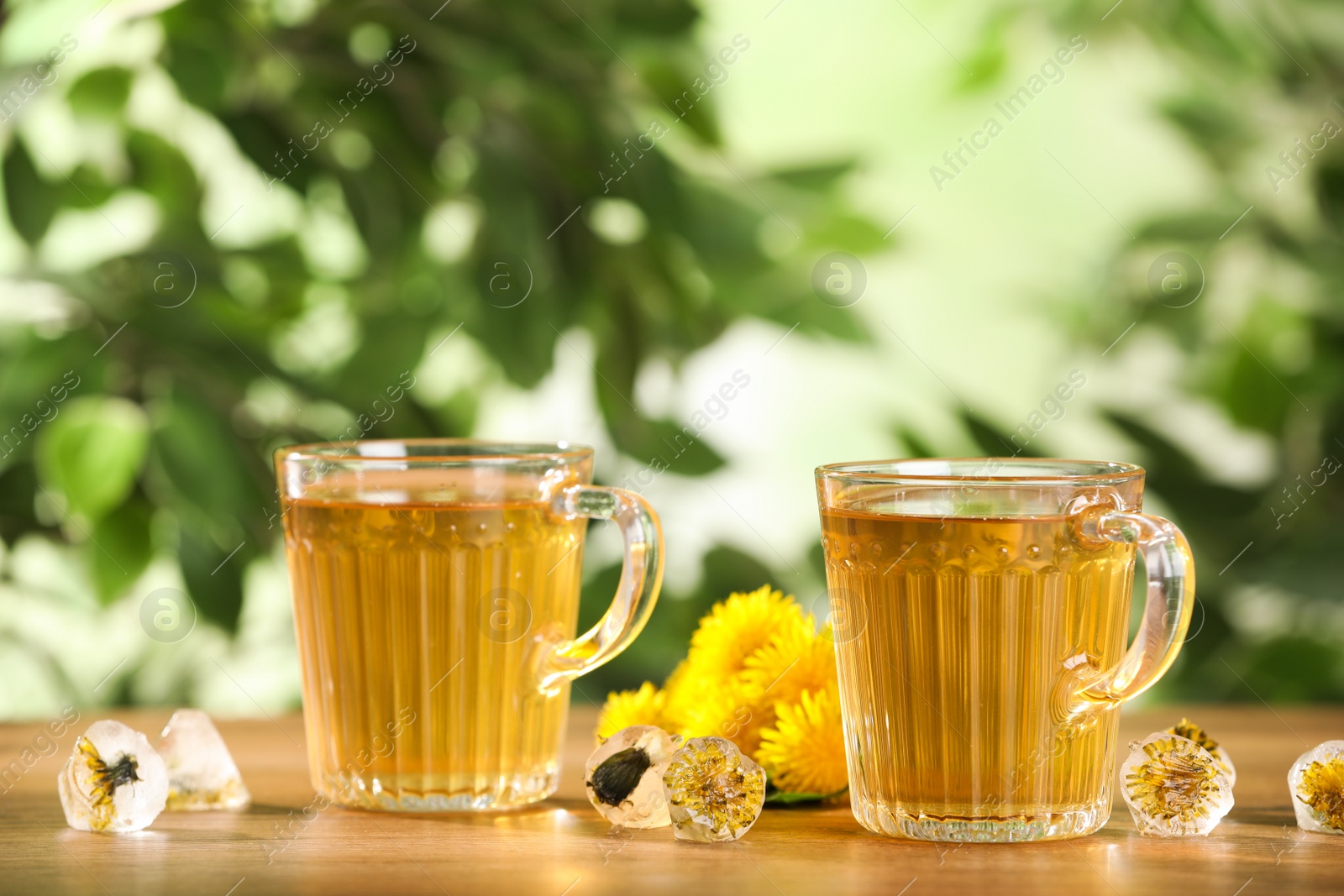 Photo of Delicious fresh tea, dandelion flowers and ice cubes on wooden table against blurred background