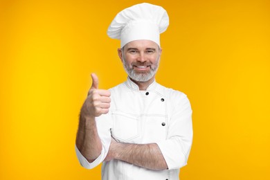 Photo of Happy chef in uniform showing thumbs up on orange background