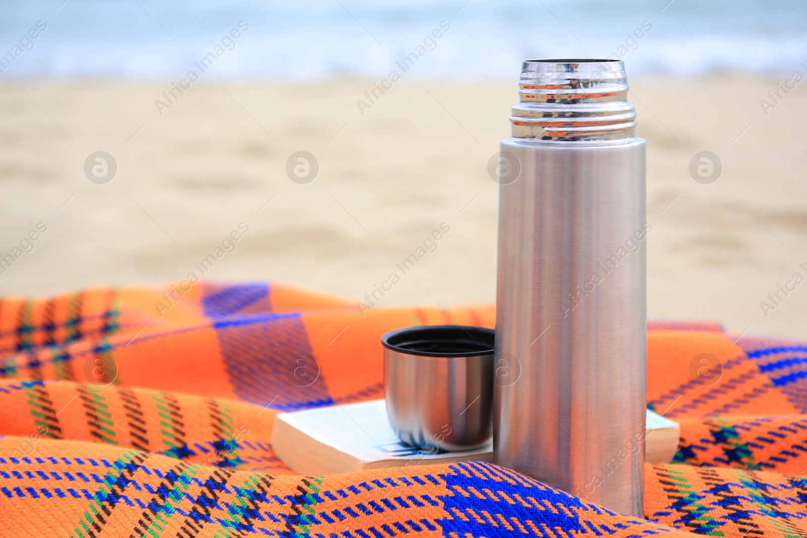 Photo of Metallic thermos with hot drink, open book and plaid on sandy beach near sea, closeup. Space for text