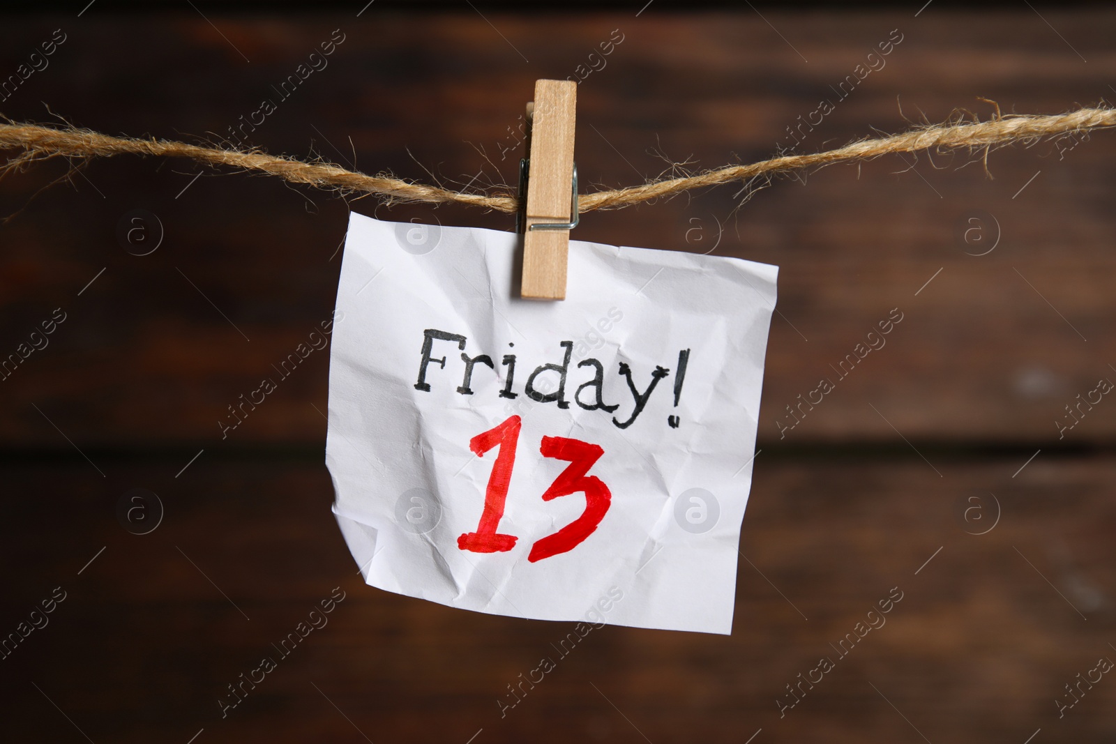 Photo of Crumpled paper note with phrase Friday! 13 hanging on twine against wooden background. Bad luck superstition