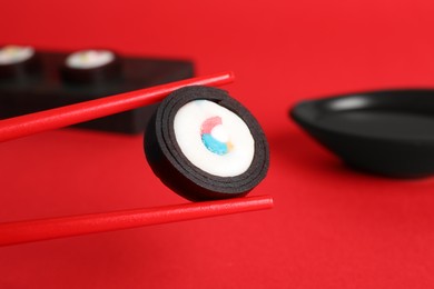 Photo of Holding sweet gummy sushi roll with chopsticks at red table, closeup. April Fools' Day