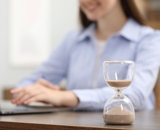 Photo of Hourglass with flowing sand on desk. Woman using laptop indoors, selective focus