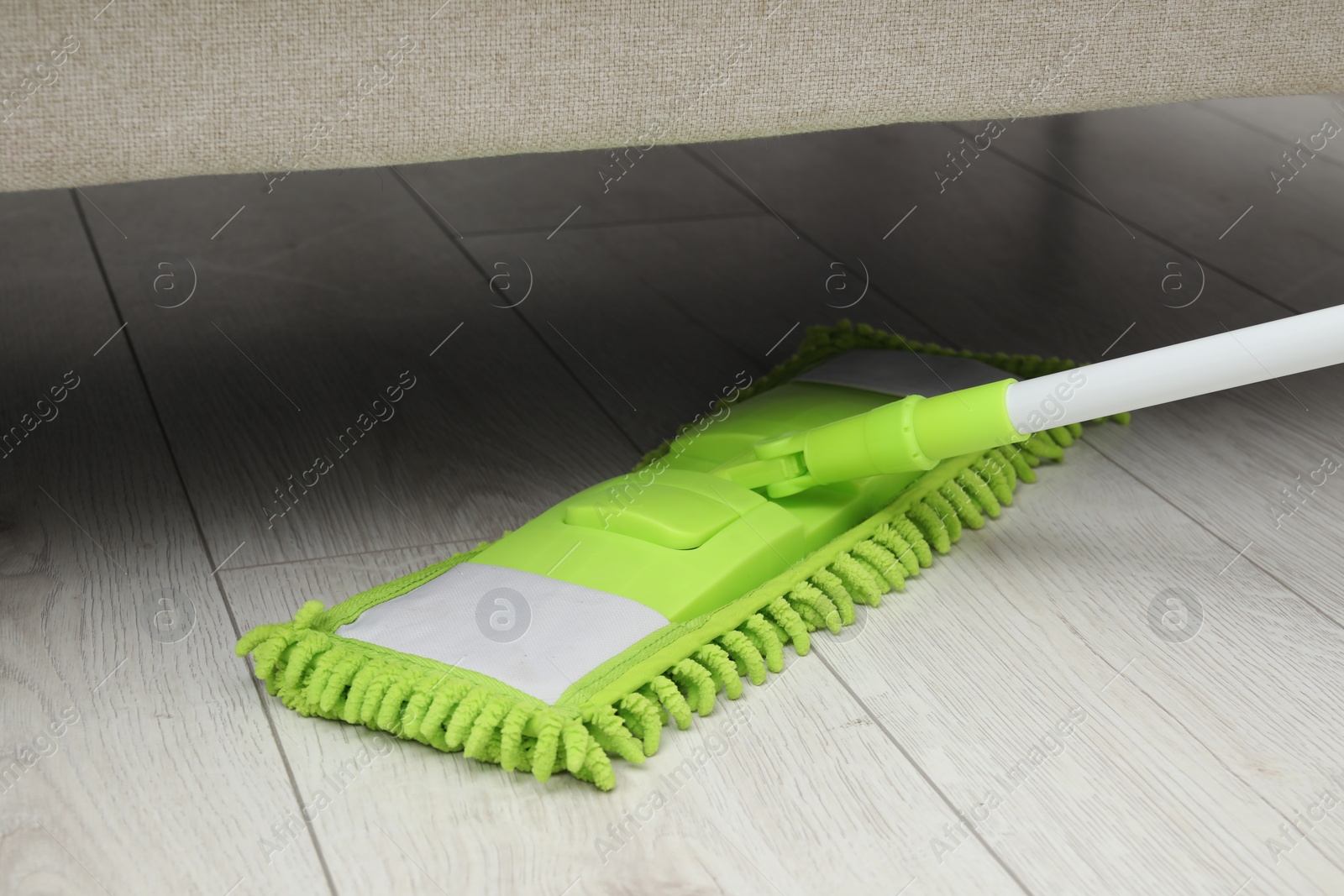 Photo of Cleaning white parquet floor under sofa with mop, closeup