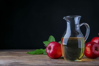 Photo of Jug of tasty juice and fresh red apples on wooden table against black background, space for text