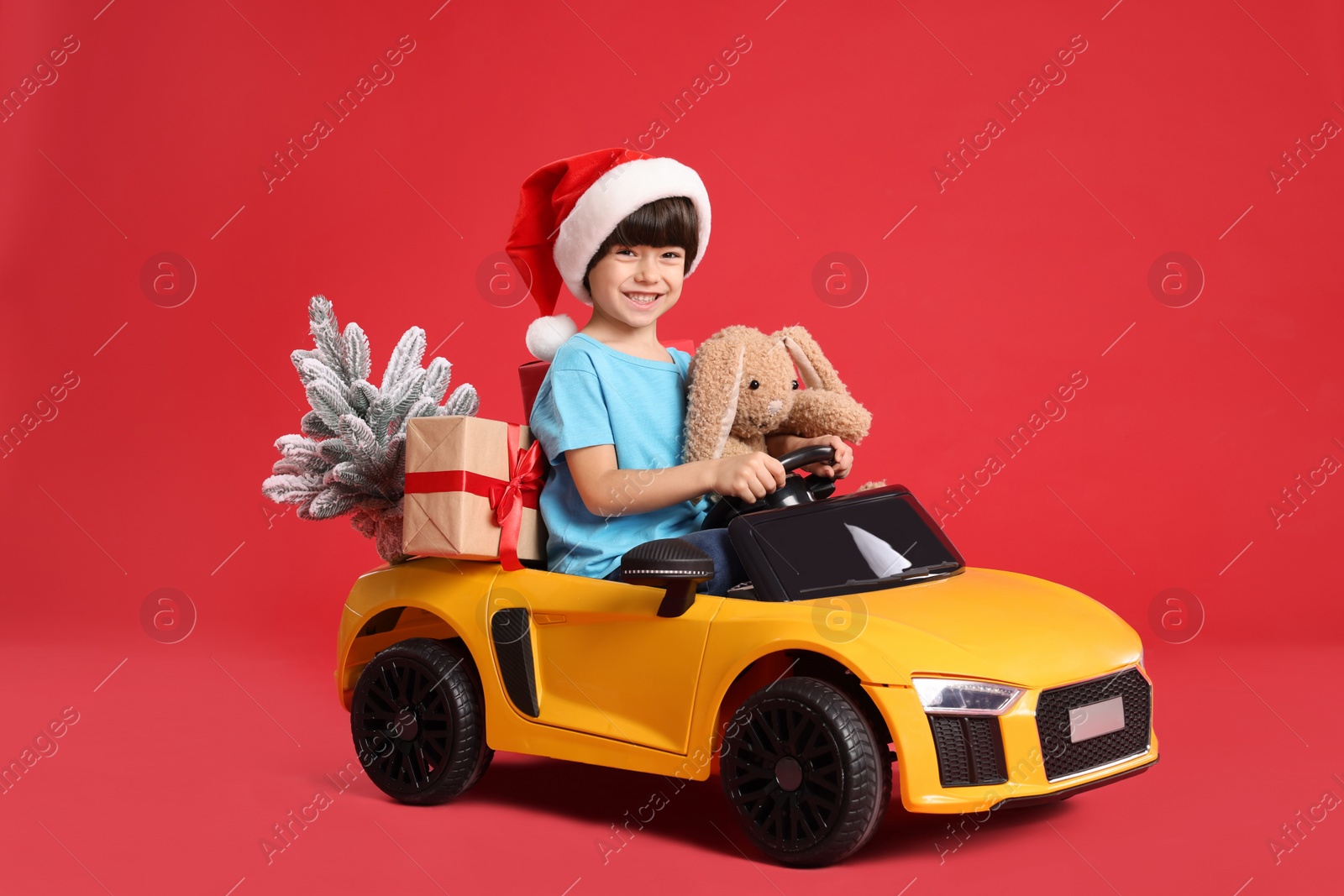 Photo of Cute little boy in Santa hat with Christmas tree, bunny toy and gift box driving children's car on red background