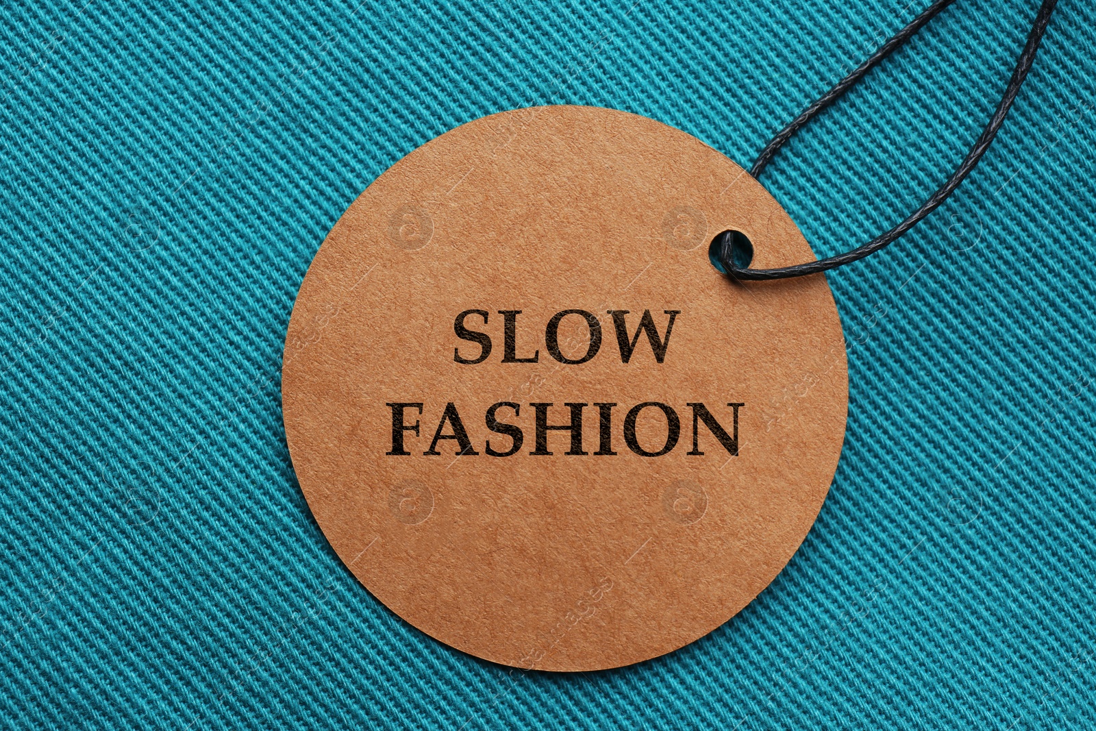 Image of Conscious consumption. Tag with words Slow Fashion on dark turquoise fabric, top view