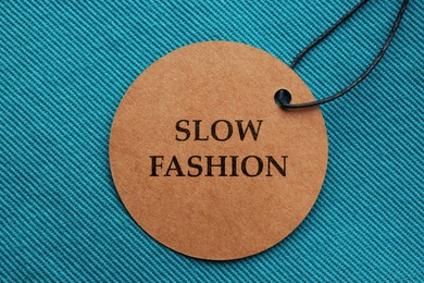 Image of Conscious consumption. Tag with words Slow Fashion on dark turquoise fabric, top view