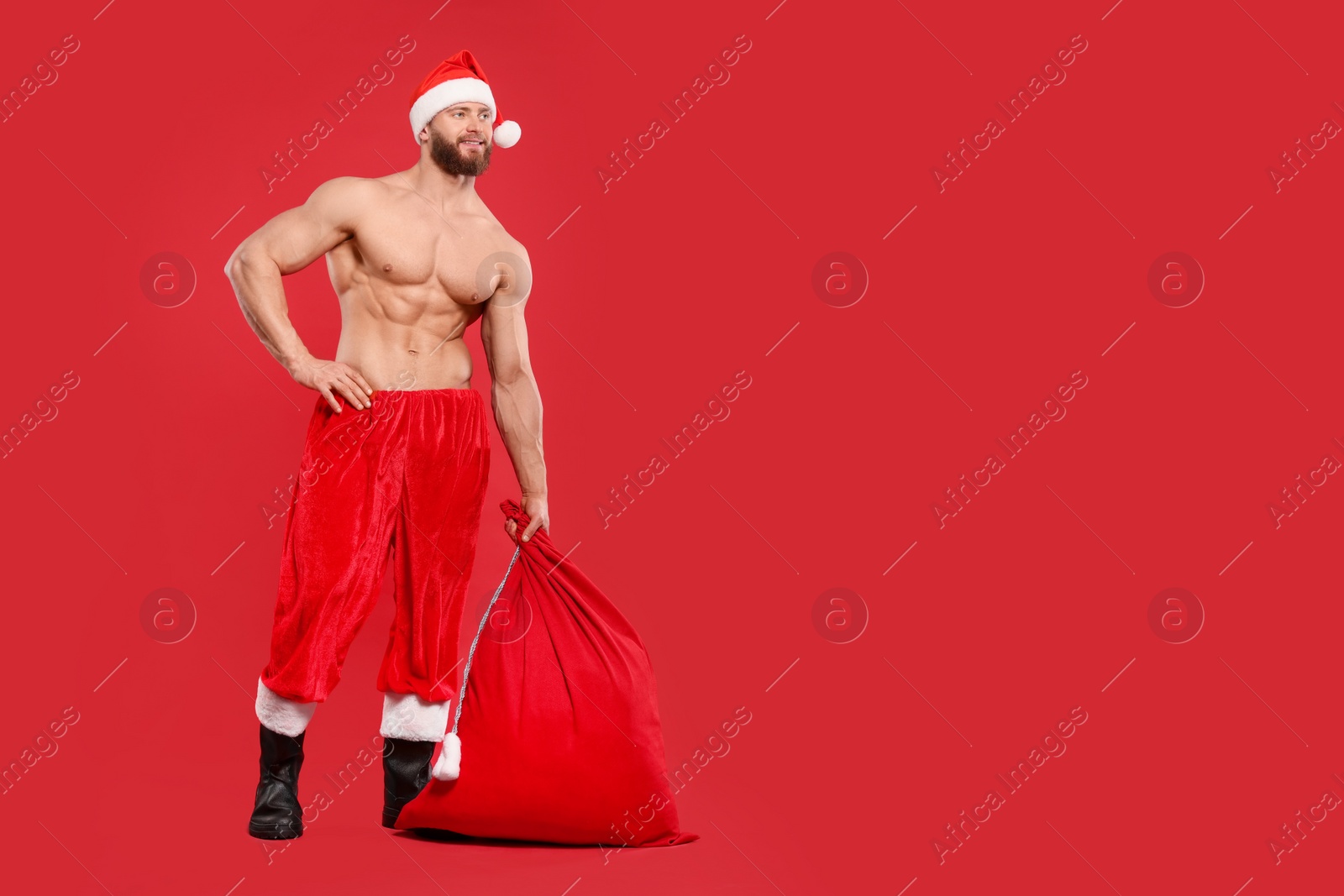 Photo of Muscular young man in Santa hat holding bag with presents on red background, space for text