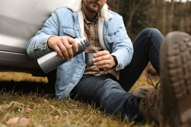 Man pouring hot drink from metallic thermos into cup lid near car outdoors, closeup