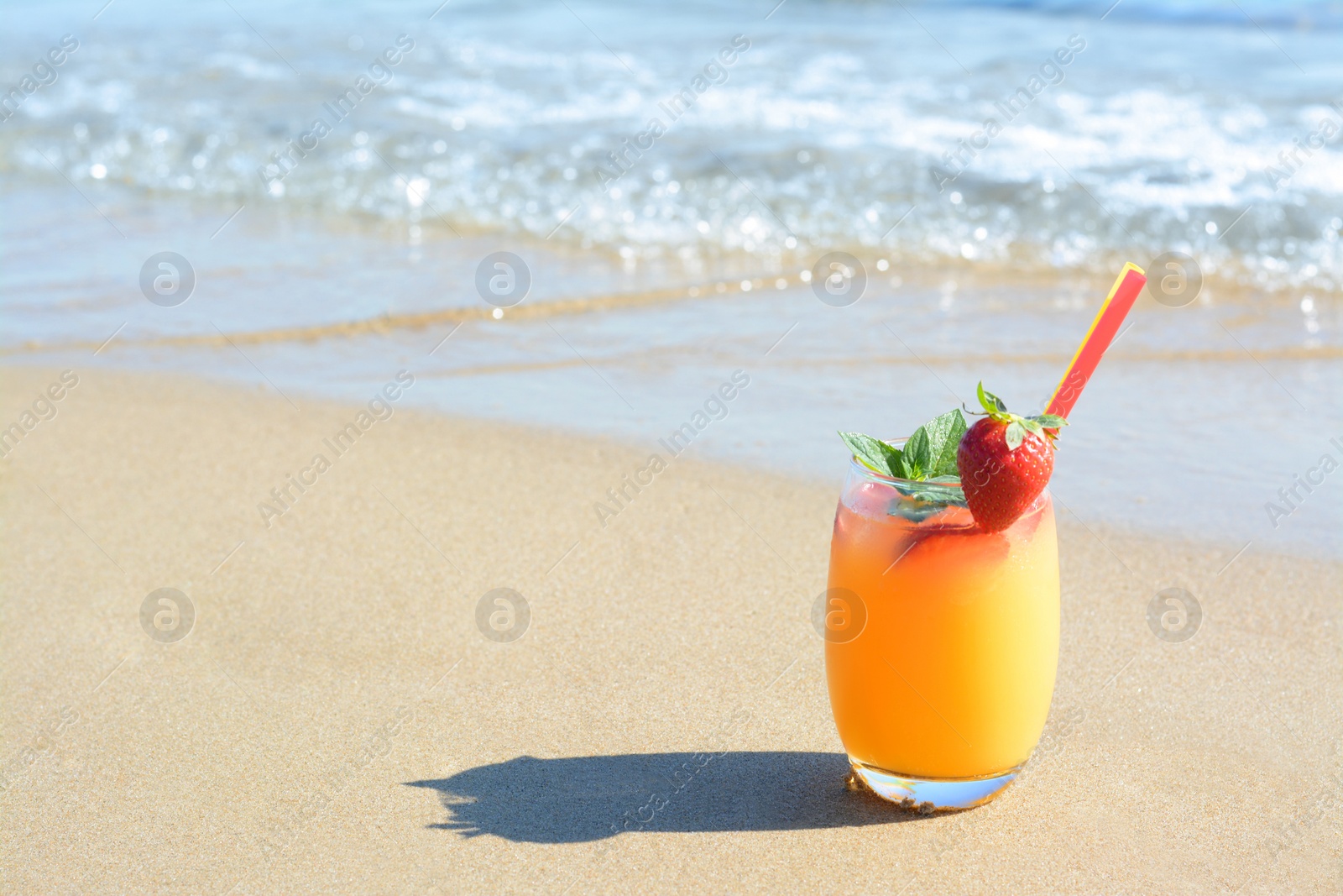 Photo of Glass of refreshing drink with strawberry on sandy beach near sea, space for text