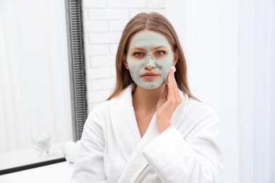 Beautiful woman removing homemade clay mask from her face indoors