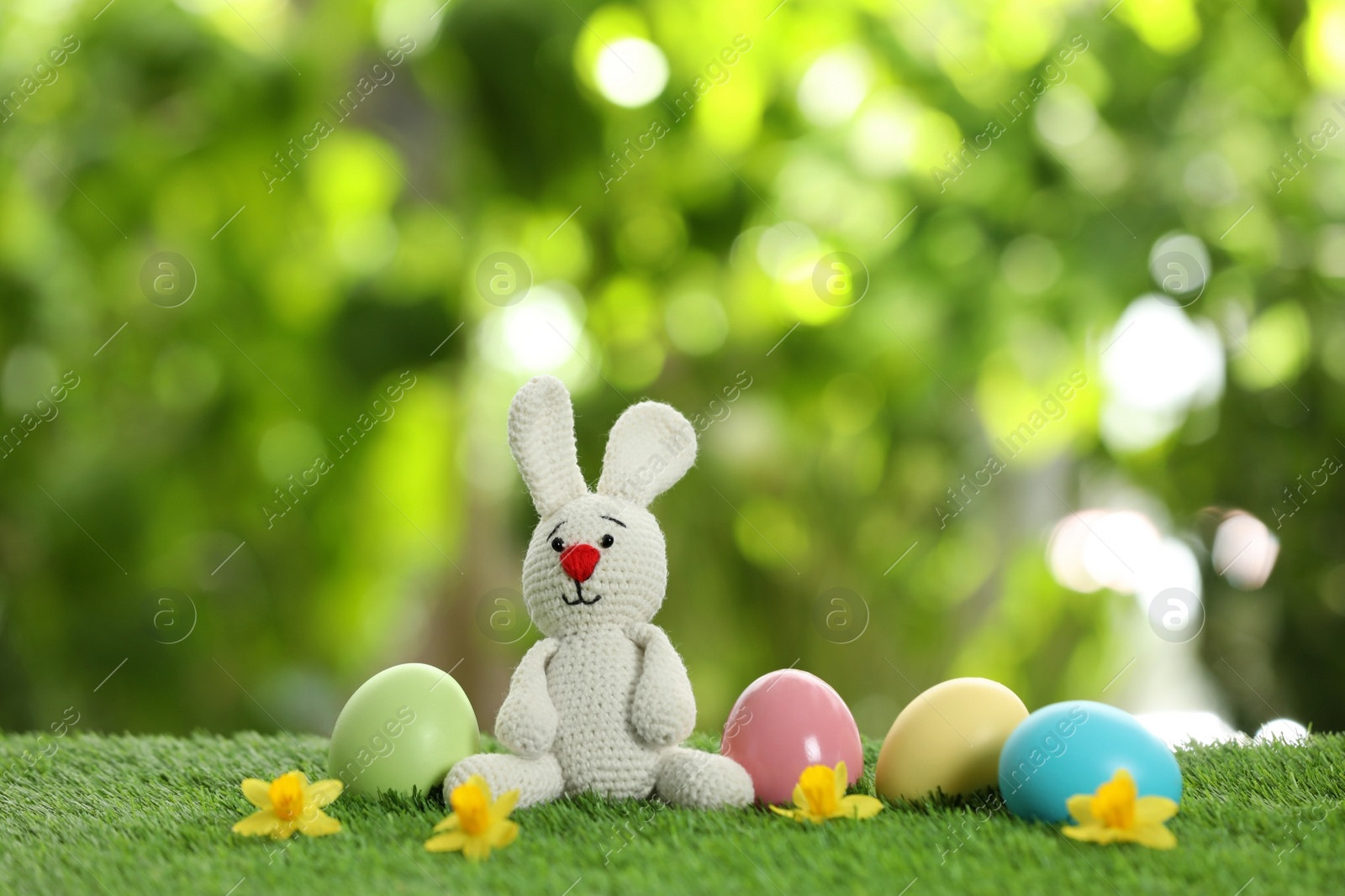 Photo of Cute Easter bunny toy and dyed eggs on green grass against blurred background, space for text