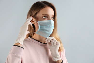 Photo of Young woman in medical gloves putting on protective mask against grey background