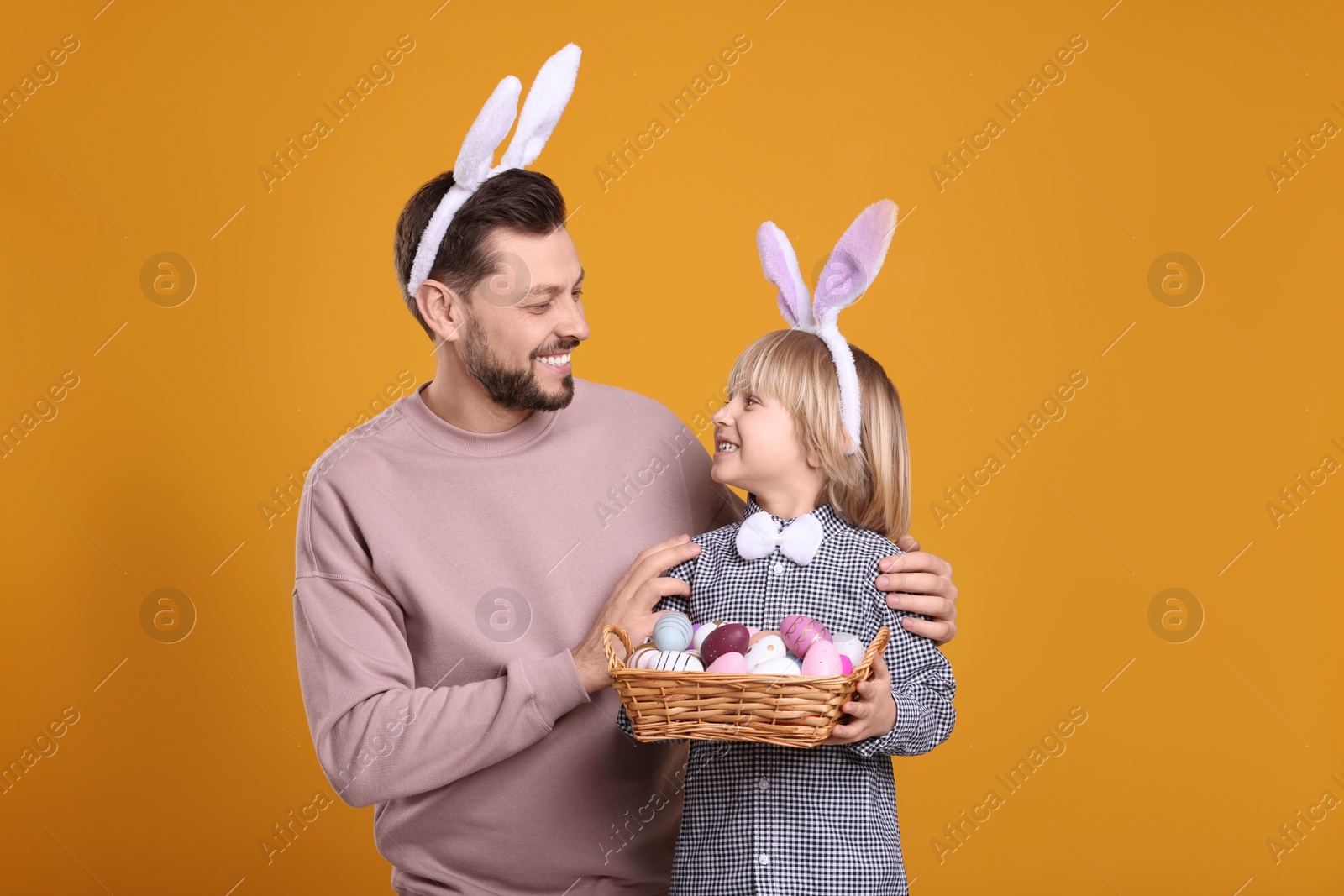 Photo of Father and son in bunny ears headbands with wicker basket of painted Easter eggs on orange background