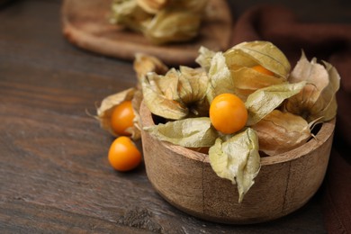 Ripe physalis fruits with calyxes in bowl on wooden table, space for text