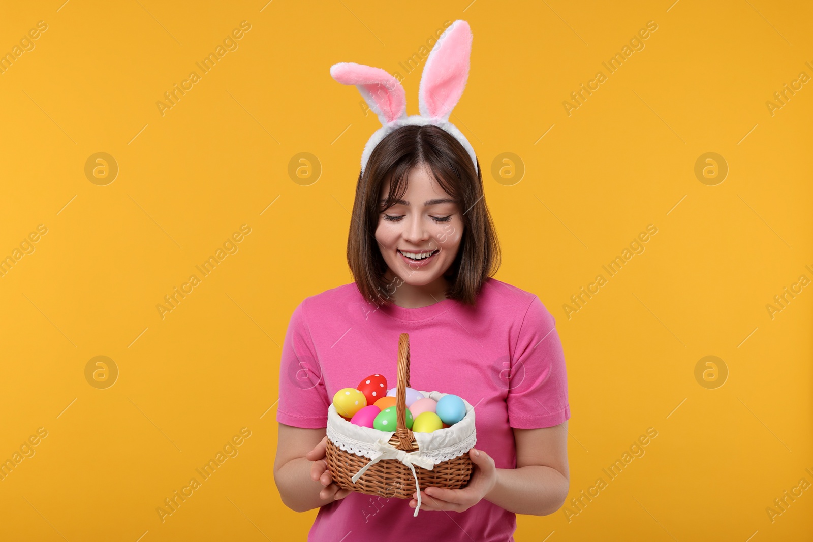 Photo of Easter celebration. Happy woman with bunny ears and wicker basket full of painted eggs on orange background