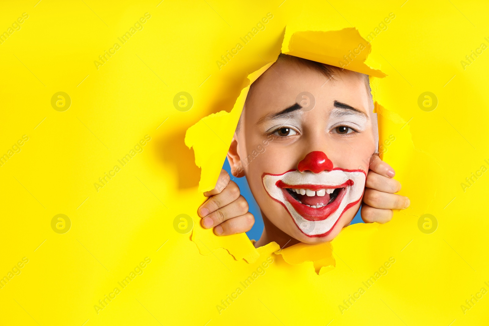 Photo of Preteen boy with clown makeup looking out of hole in yellow paper, space for text. April fool's day