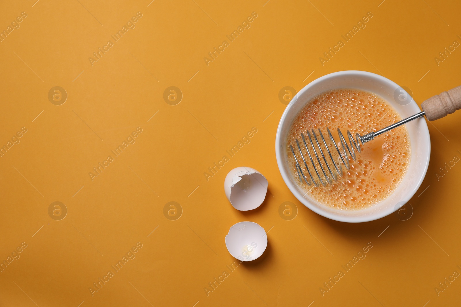 Photo of Beaten eggs, metal whisk in bowl and shells on orange background, flat lay. Space for text