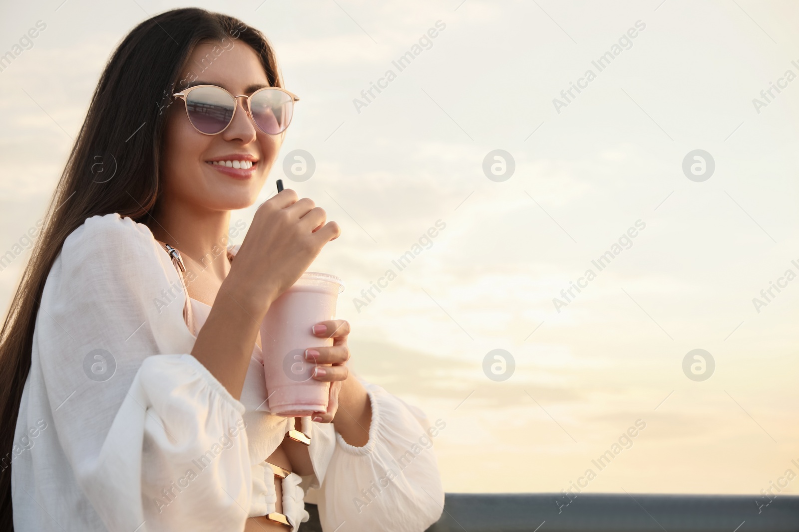 Photo of Beautiful young woman with tasty milk shake outdoors, space for text