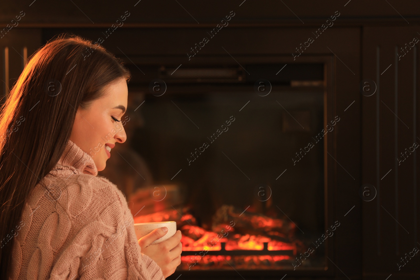 Photo of Young woman with cup of hot drink near fireplace indoors. Cozy atmosphere