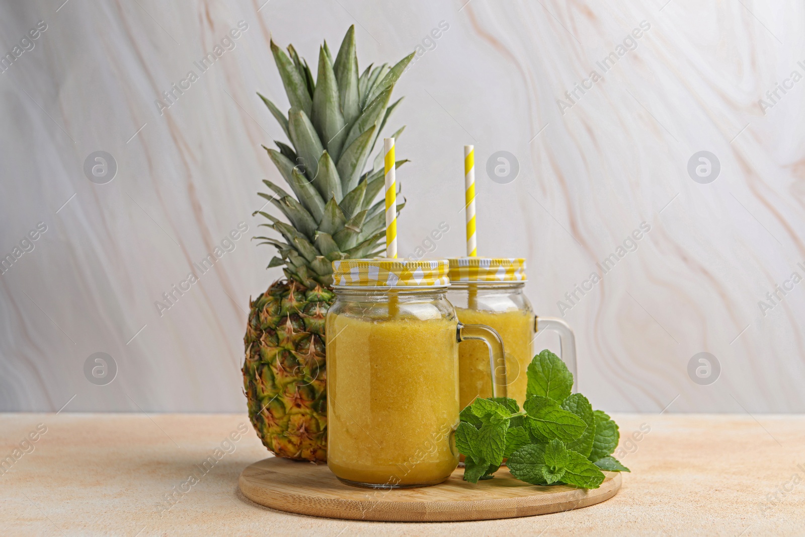 Photo of Tasty pineapple smoothie in mason jars, mint and fruit on beige table against marble background