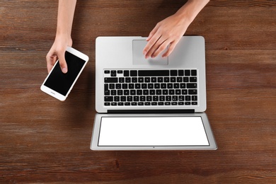 Young woman with mobile phone using laptop at wooden table, top view