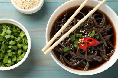 Photo of Tasty buckwheat noodle (soba) soup with chili pepper, green onion, sesame and chopsticks on light blue wooden table, flat lay