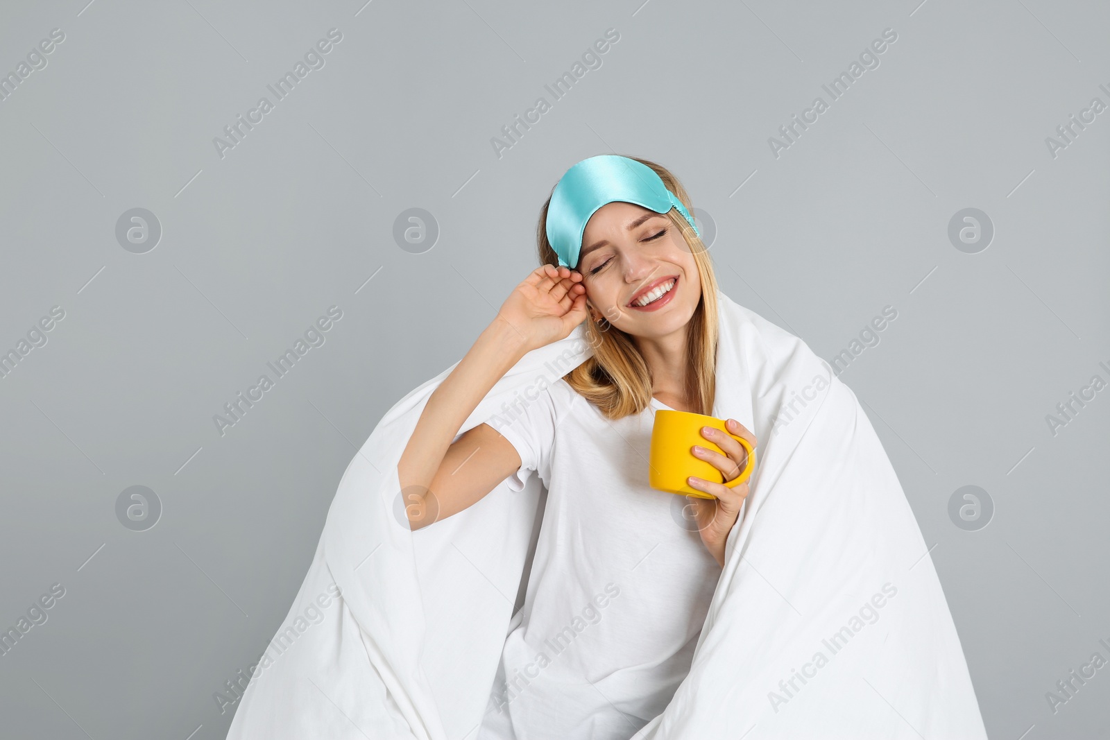 Photo of Young woman in sleeping mask wrapped with blanket holding cup on grey background
