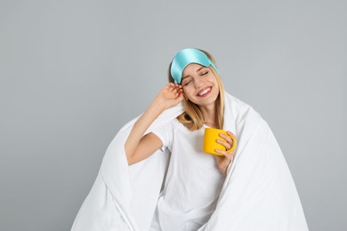 Young woman in sleeping mask wrapped with blanket holding cup on grey background