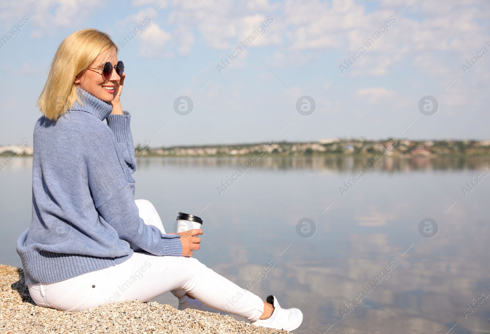 Photo of Happy woman in stylish sweater sitting on beach with cup of coffee