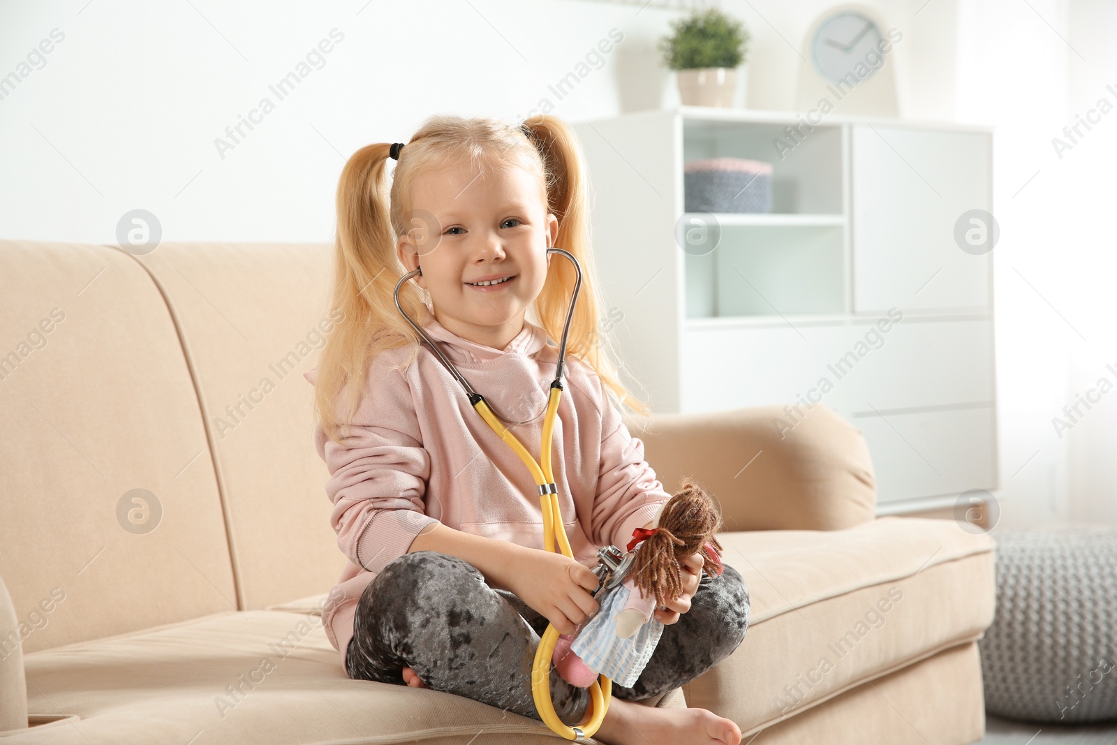 Photo of Cute child imagining herself as doctor while playing with stethoscope and doll on couch at home