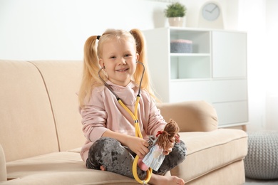 Photo of Cute child imagining herself as doctor while playing with stethoscope and doll on couch at home