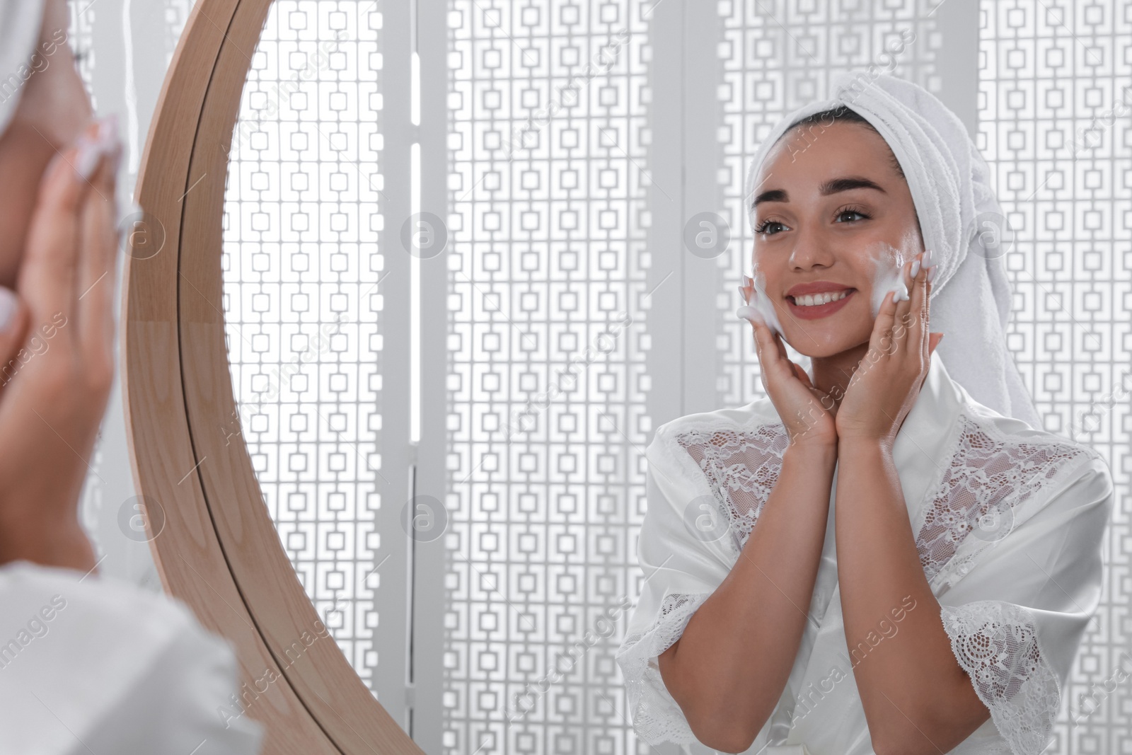 Photo of Beautiful young woman applying cleansing foam onto face near mirror in bathroom. Skin care cosmetic