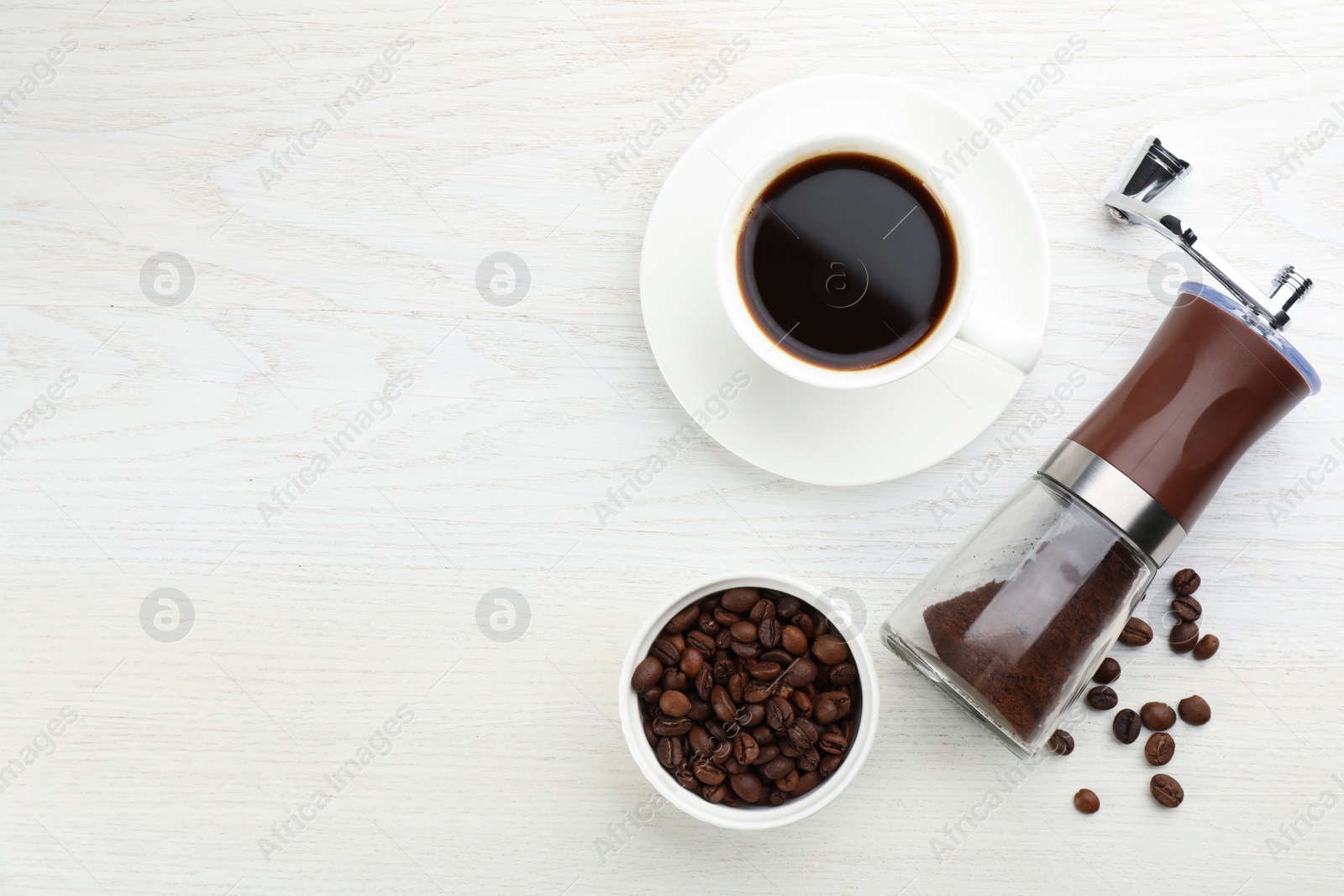 Photo of Modern manual coffee grinder with powder, beans and cup of aromatic drink on white wooden table, flat lay. Space for text