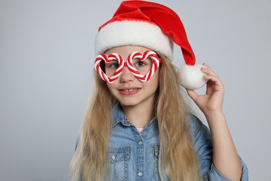 Girl wearing decorative Christmas eyeglasses in shape of hearts and Santa hat on light grey background