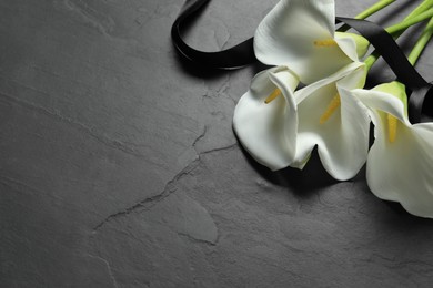 Photo of Beautiful calla lily flowers and ribbon on black table, space for text. Funeral symbols