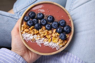 Photo of Woman holding bowl of delicious fruit smoothie with fresh blueberries, granola and coconut flakes, top view