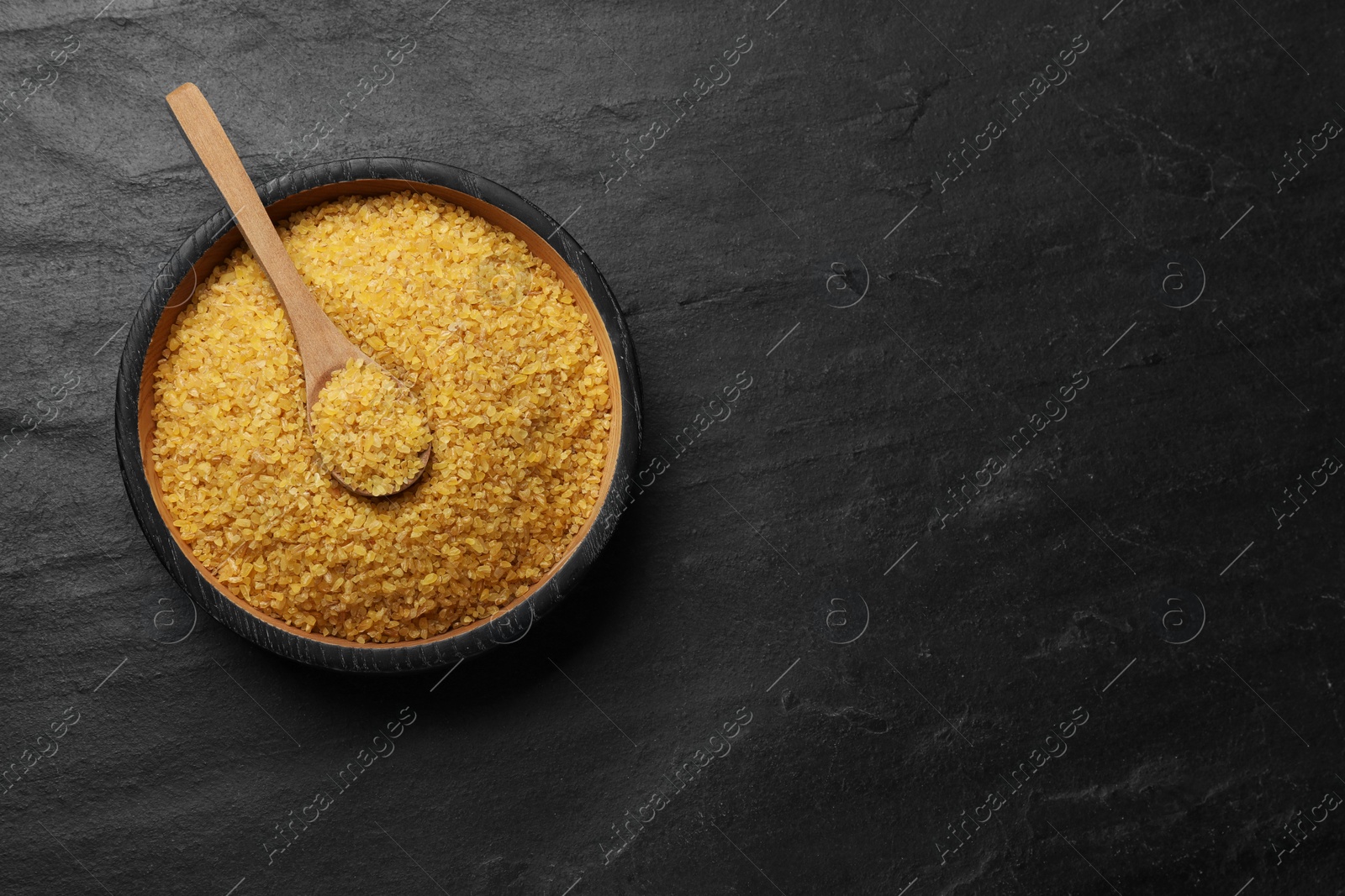 Photo of Bowl and spoon with raw bulgur on dark gray table, top view. Space for text