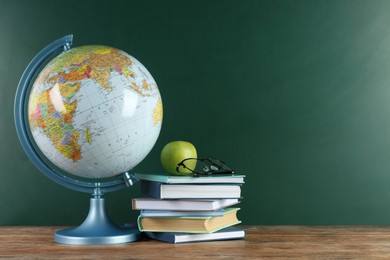 Photo of Globe, books, eyeglasses and apple on wooden table near green chalkboard, space for text. Geography lesson