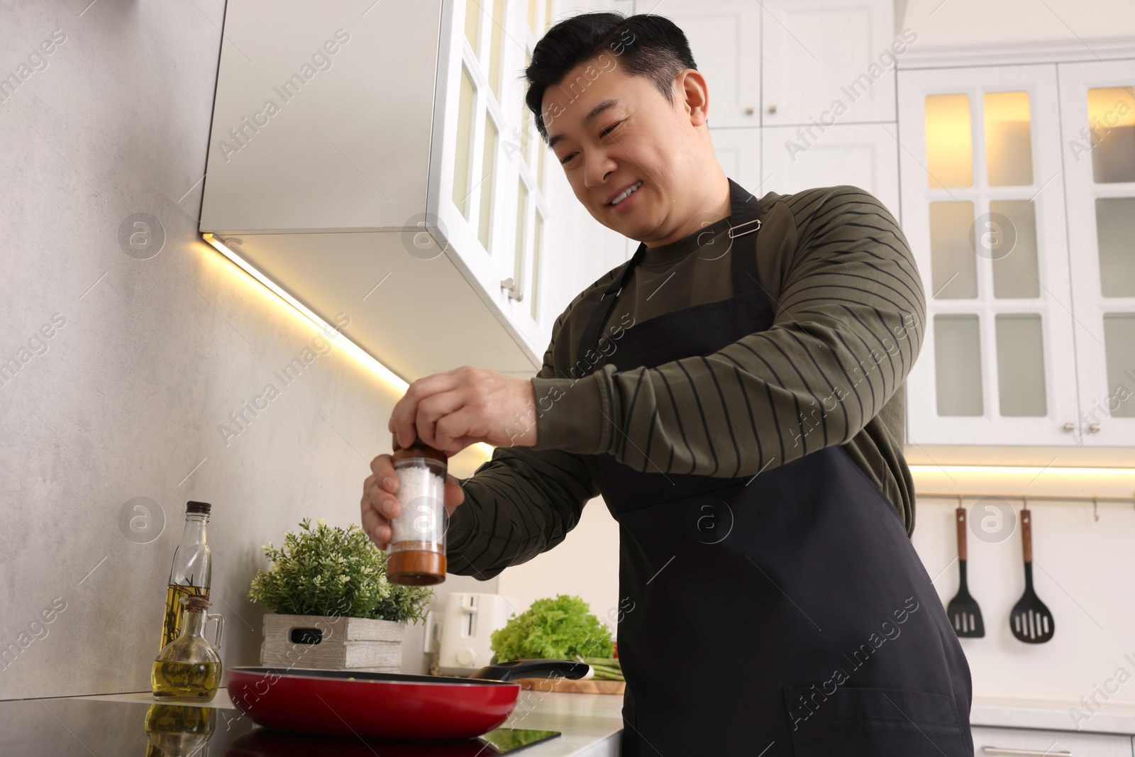 Photo of Cooking process. Man adding salt into frying pan in kitchen, low angle view