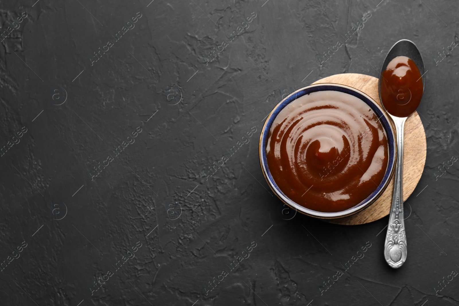 Photo of Tasty barbeque sauce in bowl and spoon on dark textured table, top view. Space for text