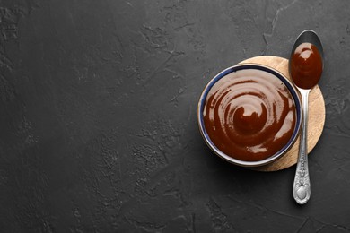 Photo of Tasty barbeque sauce in bowl and spoon on dark textured table, top view. Space for text