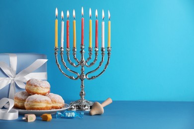 Photo of Hanukkah celebration. Menorah with burning candles, dreidels, donuts and gift box on light blue table, space for text