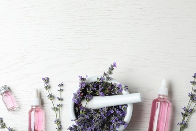 Bottles of essential oil, mortar and pestle with lavender flowers on white wooden background, flat lay. Space for text