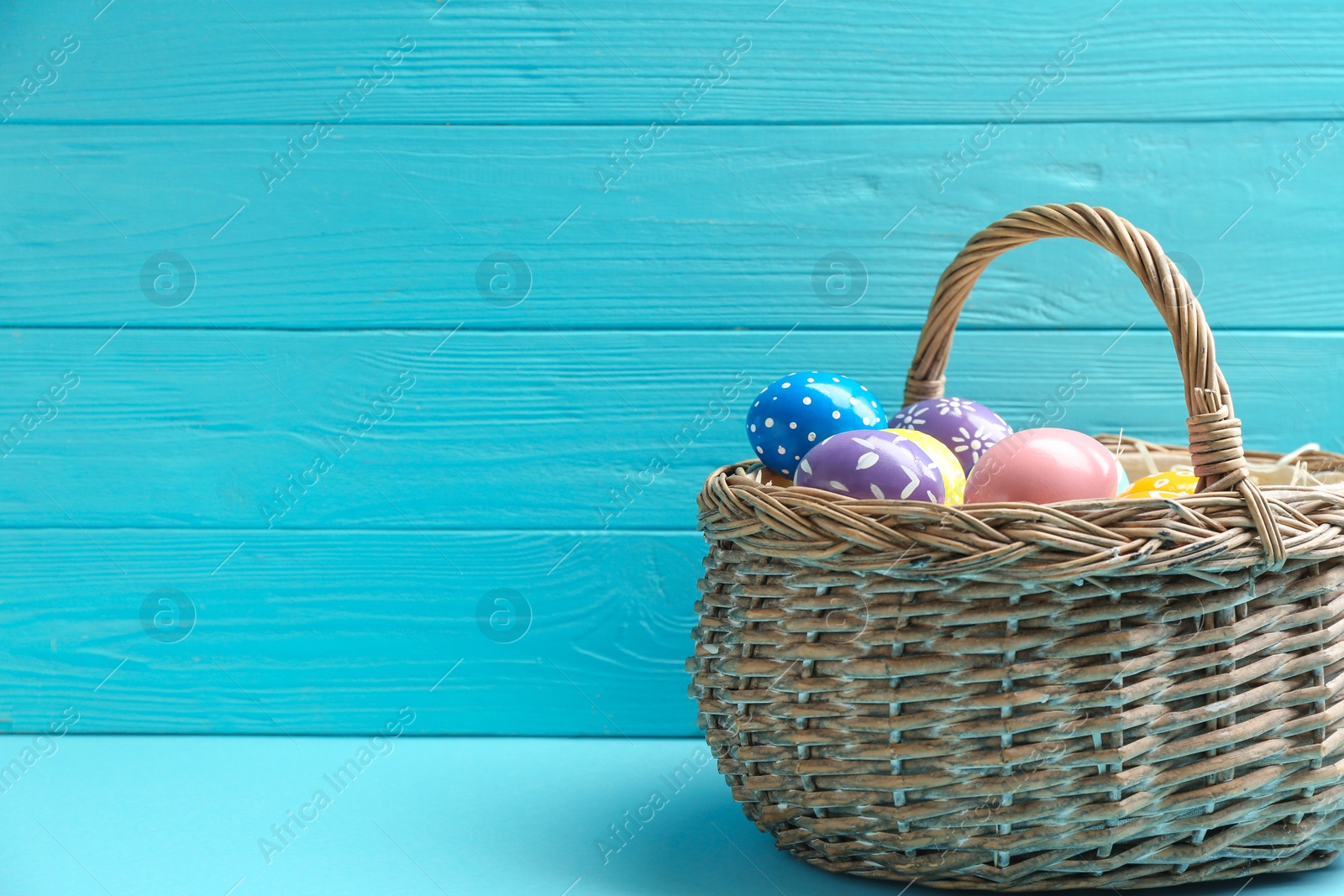 Photo of Wicker basket with painted Easter eggs on table against wooden background, space for text